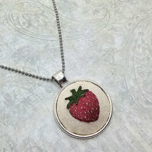 Side-angle view of a handmade strawberry embroidery necklace displayed on a light patterned background.