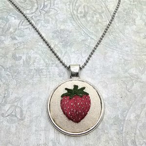 Angled view of a hand-embroidered strawberry pendant with green leaves and white seed details.