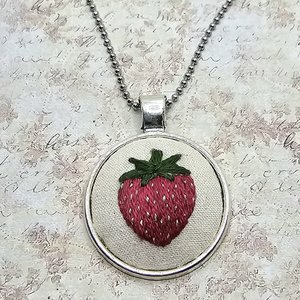 Full view of a silver-tone ball-chain necklace featuring a round hand-embroidered strawberry pendant.