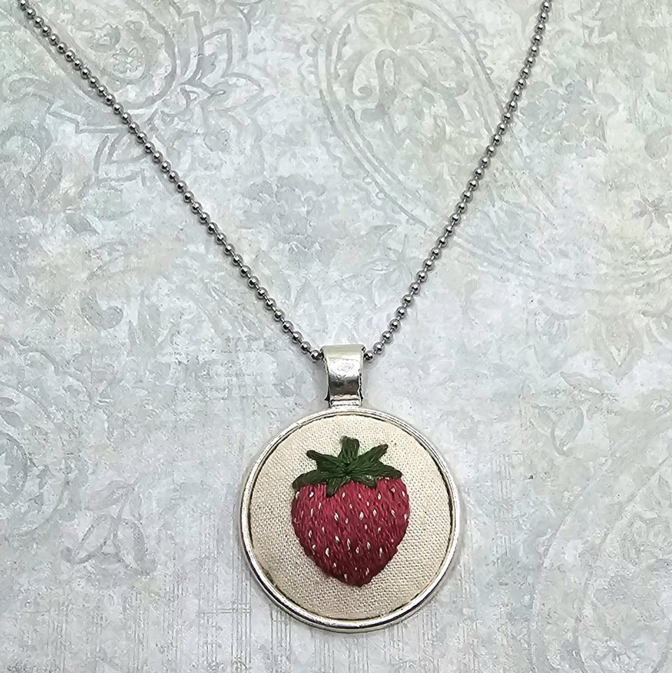 Angled close-up of a hand-embroidered strawberry pendant necklace on a silver-tone ball chain.