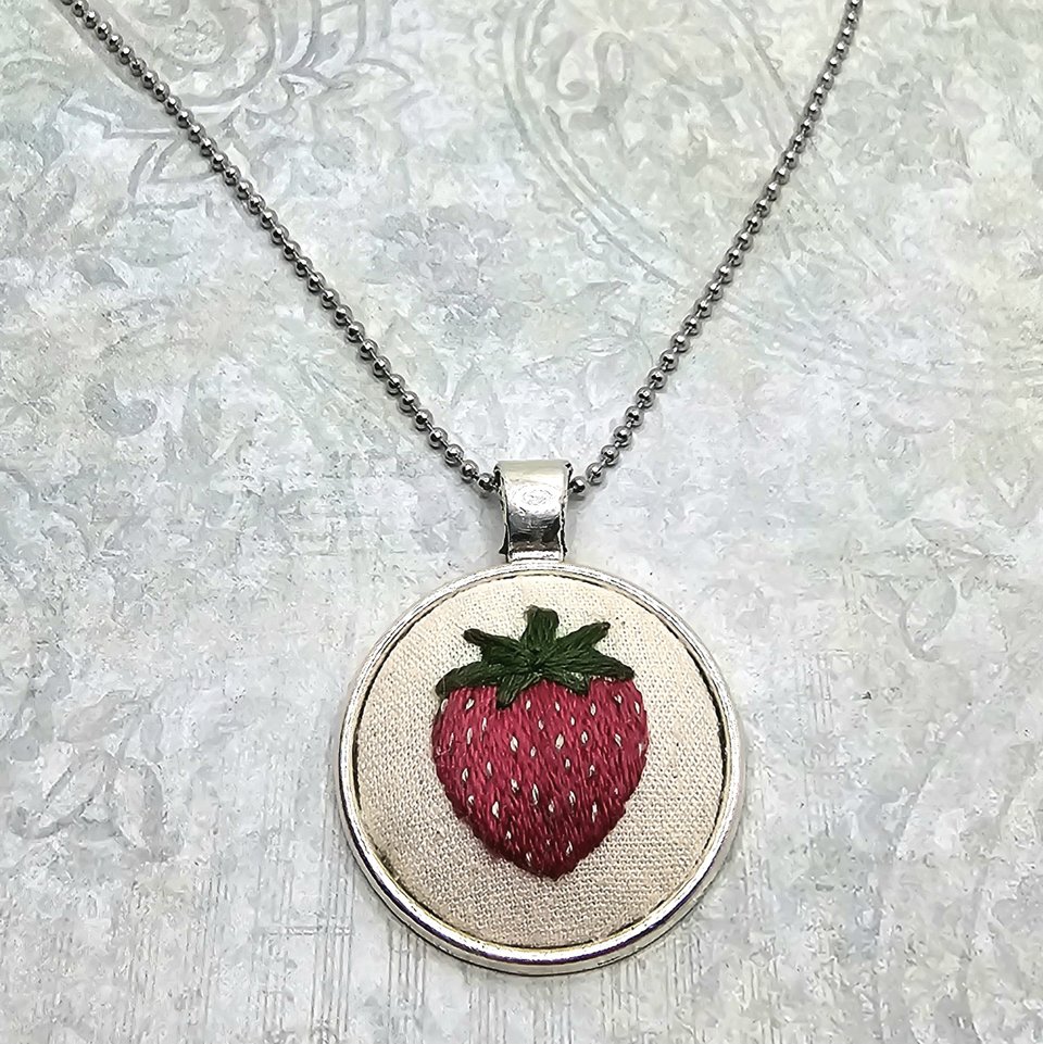 Angled view of a hand-embroidered strawberry pendant with green leaves and white seed details.