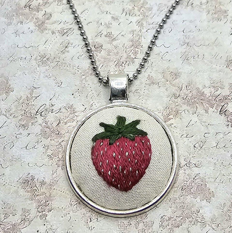 Full view of a silver-tone ball-chain necklace featuring a round hand-embroidered strawberry pendant.