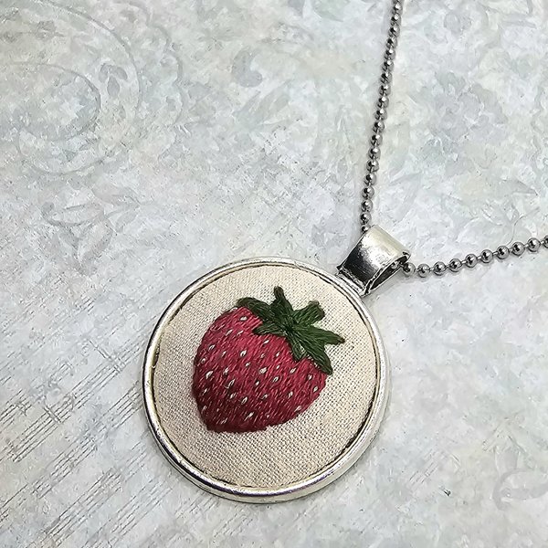 Close-up of a hand-embroidered strawberry necklace with green leaves and white seed details.